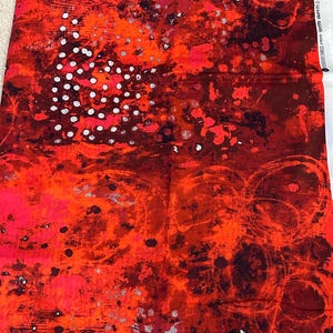 May include: Red fabric with an abstract design. The fabric features a blend of red, orange, and black hues with white and silver accents. The pattern includes splatters, dots, and circular shapes, creating a dynamic visual texture.
