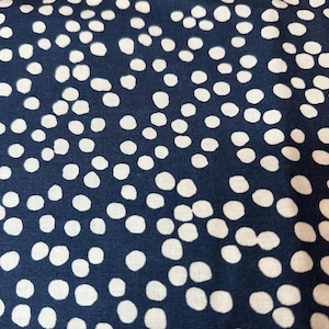 May include: Close-up of a navy blue fabric with a pattern of white, irregular polka dots. The dots are scattered across the surface, creating a textured appearance. The fabric appears to be cotton or a similar material, suitable for various sewing projects.