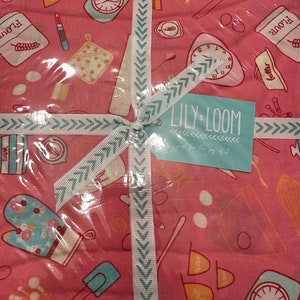 May include: Pink fabric with a white and teal chevron ribbon tied around it. The fabric has a repeating pattern of kitchen items, including a clock, measuring cups, a whisk, a rolling pin, a spatula, a mixing bowl, a sugar bag, a flour bag, and a butter dish. The fabric is labeled "Lily & Loom - Just for the joy of it."