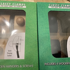 May include: Two green boxes with clear plastic windows showing wooden and black hangers. The boxes have the text "Classy Clamps The Easy Way To Display" and "Includes 4 Wooden Hangers" and "4 Wooden Hangers & Screws".