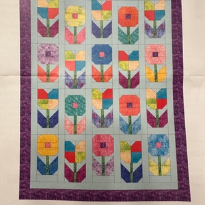 May include: A quilt pattern featuring a repeating design of colorful flowers with green stems and leaves. The quilt is surrounded by a purple border and the word "Boundless" is printed below the quilt.