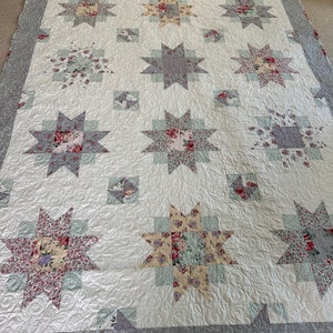 May include: A white quilt with a gray border and a floral pattern. The quilt is made up of a series of star-shaped blocks, each with a different floral fabric. The quilt is quilted with a swirling pattern.