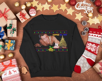 Puede incluir: Jersey navideño negro con un diseño festivo que presenta una escena de película y la frase "Yippee Ki-Yay". El jersey está rodeado de regalos envueltos, adornos y un calcetín navideño.