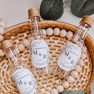 May include: Three small clear glass bottles with gold lids and personalised labels. The labels are white with a floral design and the text "SPANISH OLIVE OIL M & R 26.06.24". The bottles are sitting in a woven basket with wooden beads.