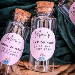 May include: Two clear glass jars with cork lids and pink labels that say "Mom's Lock of Hair" with dates. The labels have a floral design.