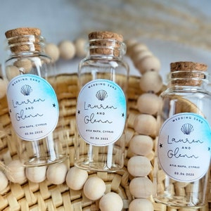 May include: Three clear glass bottles with cork tops and personalised labels. The labels are white with a blue watercolour background and a seashell graphic. The text on the labels reads "Wedding Sand", "Lauren and Glenn", "Ayia Napa, Cyprus", and "03.06.2025".