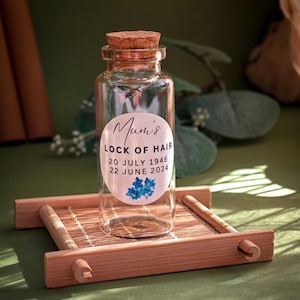 May include: A clear glass bottle with a cork stopper and a white label that reads "Mum's Lock of Hair 20 July 1946 22 June 2024". The label has a blue floral design.