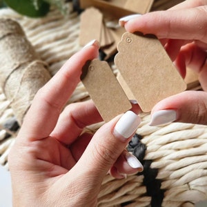 May include: Two brown paper gift tags with scalloped edges and a hole for string. The tags are held in a person's hands.