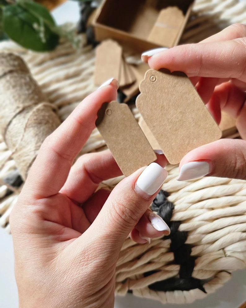 May include: Two brown paper gift tags with scalloped edges and a hole for string. The tags are held in a person's hands.