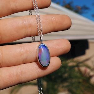 May include: A silver necklace with an oval pendant. The pendant features a colorful opal stone with hues of blue, pink, and purple. The opal is set in a silver bezel and hangs from a delicate silver chain.