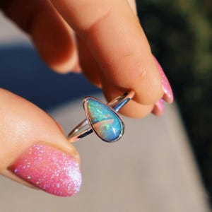 Op de afbeelding: Een zilveren ring met een peervormige opaal edelsteen. De opaal heeft blauwe, groene en witte tinten. De ring wordt tussen vingers gehouden met roze glitter nagellak.