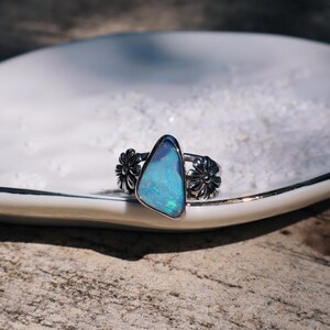 May include: A silver ring with a triangular, iridescent blue opal stone. The ring features a floral design on each side of the stone. The ring is displayed on a white, oval-shaped plate.