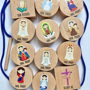 May include: Wooden religious toy set featuring illustrations of the Virgin Mary and prayers. The set includes wooden blocks and circular pieces with images and the phrases "Our Father", "Hail Mary", and "Glory Be". A blue cord is included.