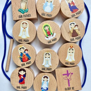May include: A set of wooden beads and blocks featuring illustrations of religious figures and symbols. The pieces include text labels such as Our Father, Hail Mary, and Glory Be. A blue cord and a wooden threading tool are included with the set.