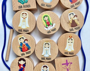 May include: A set of wooden beads and blocks featuring illustrations of religious figures and symbols. The pieces include text labels such as Our Father, Hail Mary, and Glory Be. A blue cord and a wooden threading tool are included with the set.