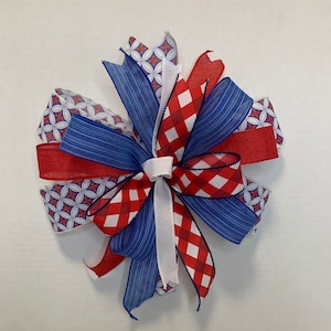 May include: A red, white, and blue bow made with patterned ribbon. The bow is tied in a knot and has a white ribbon in the center.