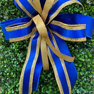 May include: A decorative bow crafted from royal blue satin ribbon with shimmering gold glitter edges. The bow features multiple loops and long, flowing tails, with a central knot securing the layered ribbon structure in a symmetrical arrangement.