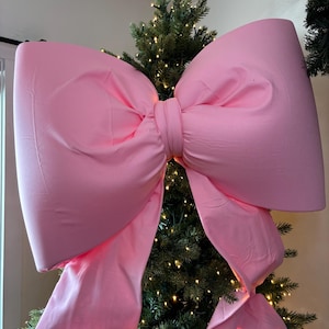 May include: A large, solid light pink fabric bow designed as a tree topper. The bow features two wide, structured loops and two long, flowing tails that drape downward. The material has a smooth, matte finish and is positioned atop a green tree.