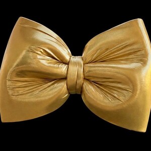 May include: A large, metallic gold bow with a smooth, reflective surface and soft, gathered folds radiating from the center knot. The bow features a structured shape with wide, pointed ends, set against a solid black background.