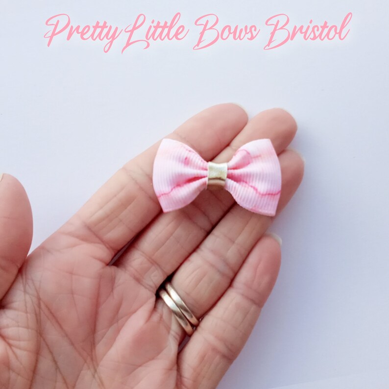May include: A small pink bow with a white marble pattern and a gold center. The bow is held in a hand.