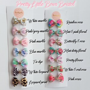 May include: A set of 10 pairs of handmade hair bows with various patterns and colors, including white marble, pink marble, blue marble, tie dye, leopard print, rainbow rose, mint and pink floral, butterfly and rose, mint dotty floral, and white and red rose. The bows are displayed on a white card with the text "Pretty Little Bows Bristol" at the top.