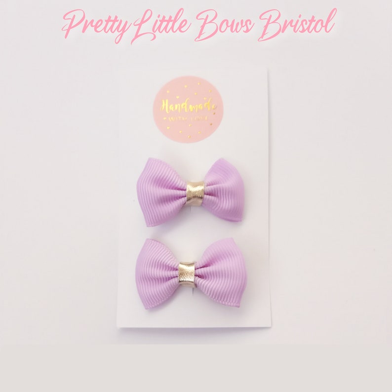 May include: Two lavender hair bows with gold accents. The bows are on a white card with the text "Handmade with love" on a pink circle.