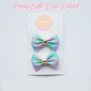 May include: Two pastel rainbow tie dye hair bows with gold accents. The bows are on a white card with the text "Handmade with Love" on a pink circle.