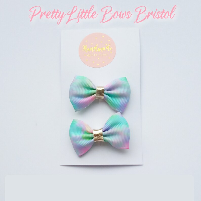 May include: Two pastel rainbow tie dye hair bows with gold accents. The bows are on a white card with the text "Handmade with Love" on a pink circle.