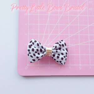 May include: A white and black spotted bow with a gold center. The bow is on a pink cutting mat with white grid lines. The text "Pretty Little Bows Bristol" is visible in the top left corner of the image.