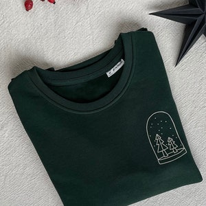 Könnte beinhalten: Ein dunkelgrünes Sweatshirt mit einem weißen Umriss eines Schneeballs mit zwei Kiefern darin. Das Sweatshirt hat ein Etikett mit der Aufschrift "#IM ORGANIC" und "M".