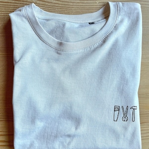 Könnte beinhalten: Ein hellgraues T-Shirt mit einem kleinen schwarzen gestickten Design aus einer Säge, einer Zange und einem Hammer mit den Buchstaben "PVT" über den Werkzeugen.