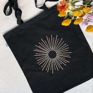 Könnte beinhalten: Schwarze Tragetasche mit einem goldenen Sonnenstrahlen-Design. Die Tasche hat lange Henkel und ist aus strapazierfähigem Canvas gefertigt.