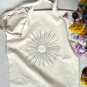 Könnte beinhalten: Eine weiße Canvas-Tragetasche mit einem schwarzen Sonnenmotiv. Die Sonne hat einen Kreis in der Mitte und Strahlen, die nach außen strahlen.
