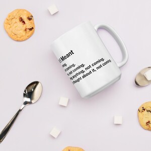 May include: White ceramic mug with the text "I Meant" followed by a list of phrases. The mug has a curved handle and is surrounded by cookies, sugar cubes, and a spoon.