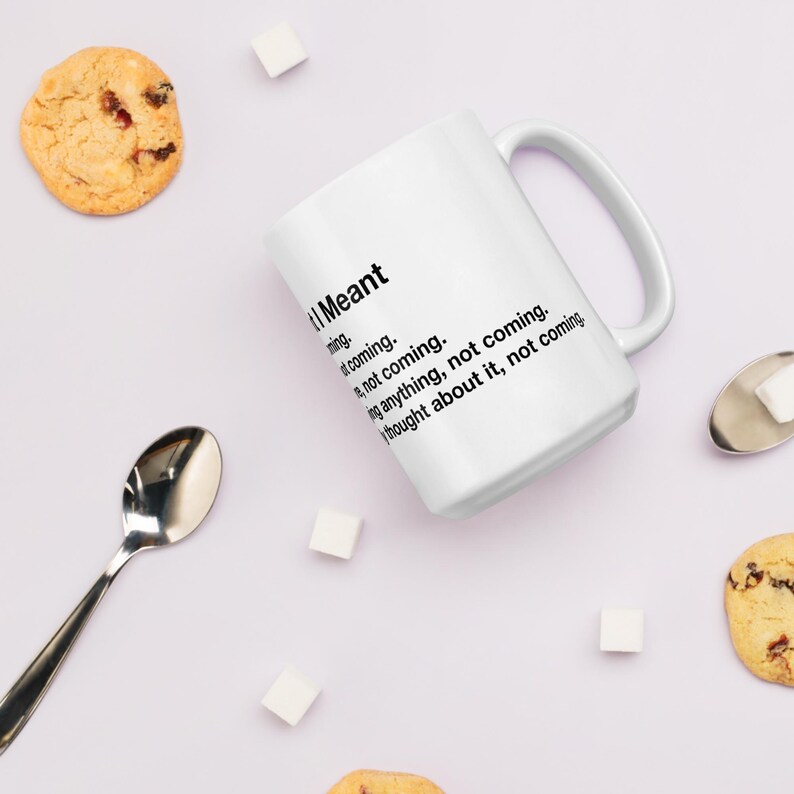 May include: White ceramic mug with the text "I Meant" followed by a list of phrases. The mug has a curved handle and is surrounded by cookies, sugar cubes, and a spoon.