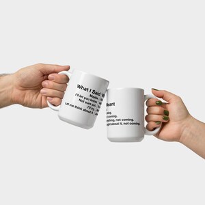 May include: Two white ceramic mugs with black text are held up in a clinking gesture. One mug reads "What I Said" with a list of phrases. The other mug reads "Meant" with a list of phrases. The mugs are held by two hands.