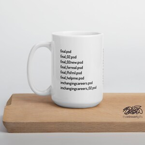 Op de afbeelding: Een witte keramische mok met een zwart ontwerp van een lijst met bestandsnamen die eindigen op .psd. De tekst op de mok luidt: "final.psd, final_02.psd, final_02new.psd, final_forreal.psd, final_ffsfml.psd, final_helpme.psd, imchangingcareers.psd, imchangingcareers_02.psd". De mok staat op een houten oppervlak. De tekst "theintrovertgifts" is zichtbaar in de rechterbenedenhoek van de afbeelding.
