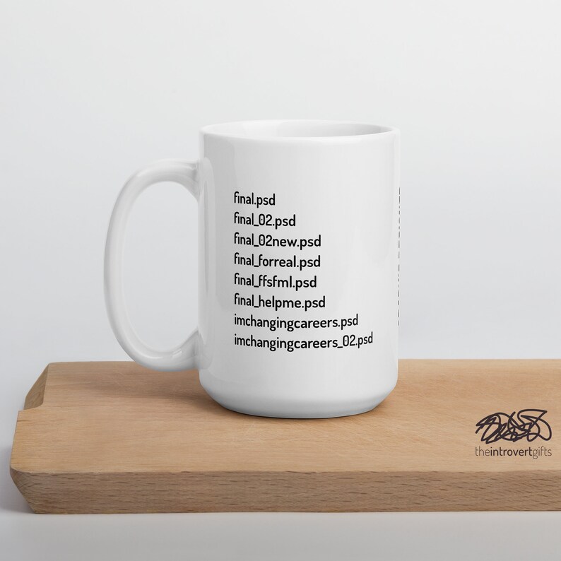 Op de afbeelding: Een witte keramische mok met een zwart ontwerp van een lijst met bestandsnamen die eindigen op .psd. De tekst op de mok luidt: "final.psd, final_02.psd, final_02new.psd, final_forreal.psd, final_ffsfml.psd, final_helpme.psd, imchangingcareers.psd, imchangingcareers_02.psd". De mok staat op een houten oppervlak. De tekst "theintrovertgifts" is zichtbaar in de rechterbenedenhoek van de afbeelding.