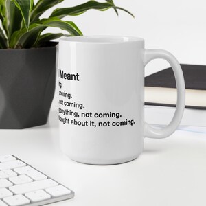 May include: White ceramic mug with a black handle. The mug features black text that reads "I Meant" followed by a list of phrases. A plant and keyboard are visible in the background.