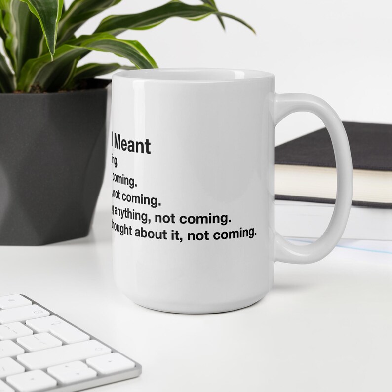 May include: White ceramic mug with a black handle. The mug features black text that reads "I Meant" followed by a list of phrases. A plant and keyboard are visible in the background.