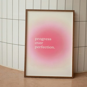 Peut inclure: Une impression encadrée avec un fond dégradé rose et le texte "progress over perfection." L'impression est appuyée contre un mur en carrelage blanc.
