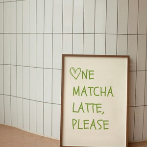May include: A framed print with the words "One Matcha Latte, Please" in green, with a green heart above "One." The print has a cream-coloured background and a brown wooden frame. The print is leaning against a white tiled wall.