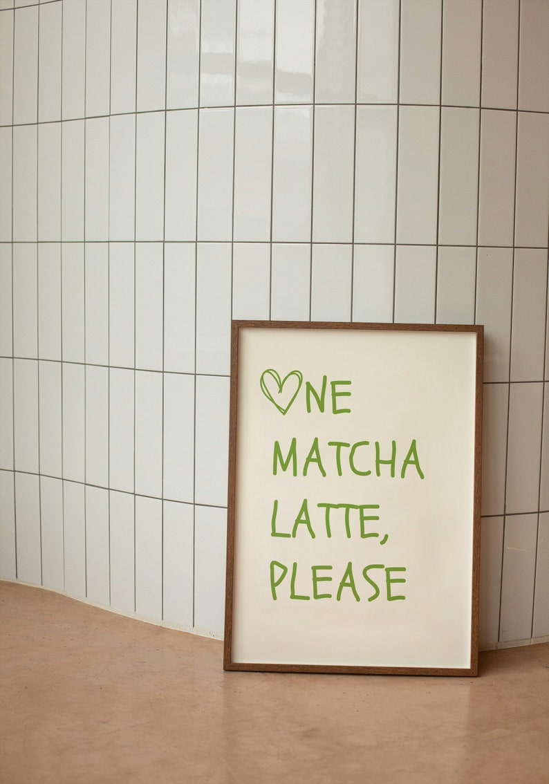 May include: A framed print with the words "One Matcha Latte, Please" in green, with a green heart above "One." The print has a cream-coloured background and a brown wooden frame. The print is leaning against a white tiled wall.