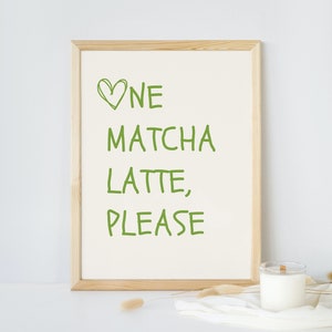 May include: A framed print with the words "One Matcha Latte, Please" in green lettering. The print is in a light wood frame and sits on a white surface. A candle in a glass jar and decorative sprigs are also on the surface.