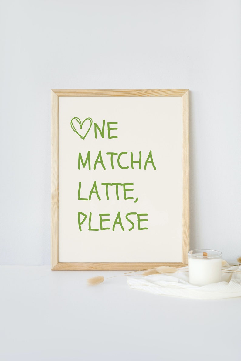 May include: A framed print with the words "One Matcha Latte, Please" in green lettering. The print is in a light wood frame and sits on a white surface. A candle in a glass jar and decorative sprigs are also on the surface.