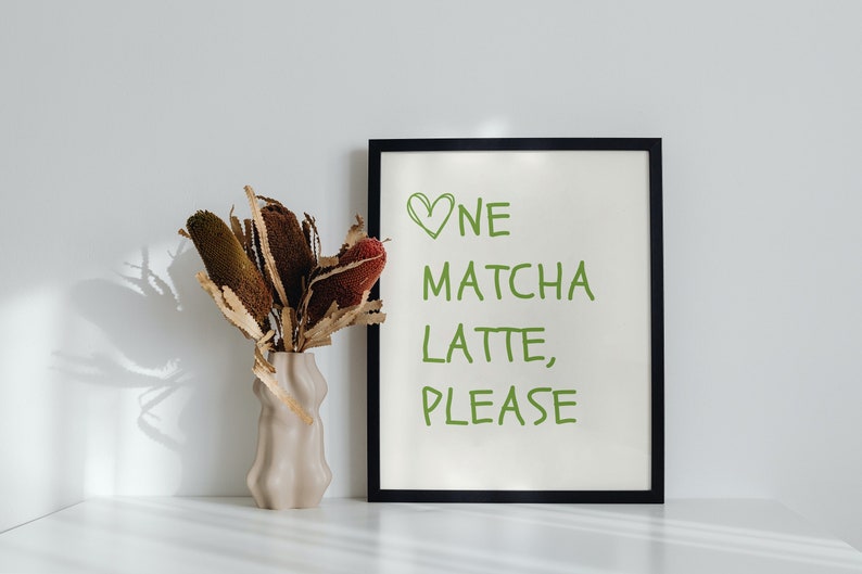 May include: A framed print with the words "One Matcha Latte, Please" in green. A beige vase with dried flowers is positioned to the left of the frame. The image is set against a white background.