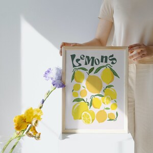 May include: A framed print with the word "Lemons" in green text and a yellow and green illustration of lemons. The print is being held by a person wearing a light beige shirt and white pants.