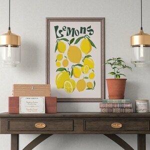 May include: A framed print with the text "Les  Mons" and a colorful illustration of lemons. The print is hanging on a white wall above a wooden table with a small wooden box, a stack of books, a potted plant, and two gold pendant lights.
