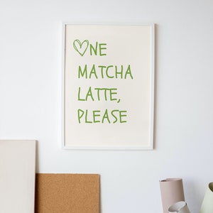 May include: A framed print with the words "One Matcha Latte, Please" in green, with a green heart symbol. The print is in a white frame and hangs on a white wall. A corkboard and a woven basket are also visible.