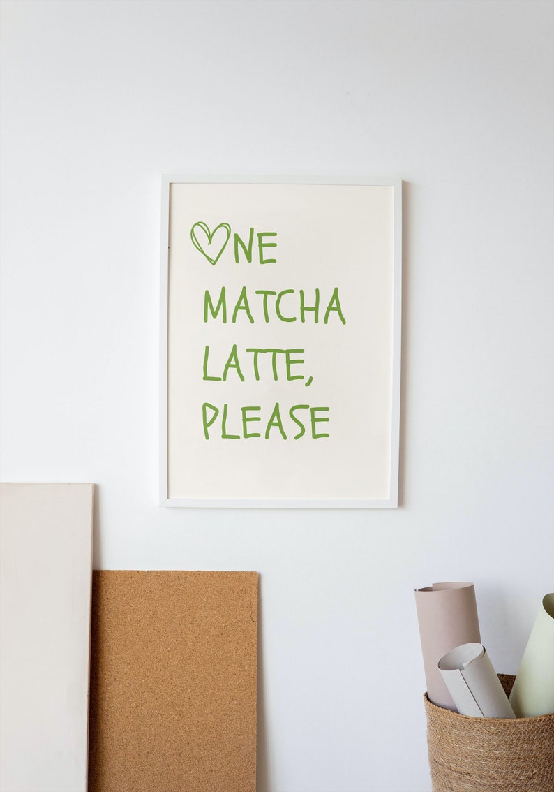 May include: A framed print with the words "One Matcha Latte, Please" in green, with a green heart symbol. The print is in a white frame and hangs on a white wall. A corkboard and a woven basket are also visible.